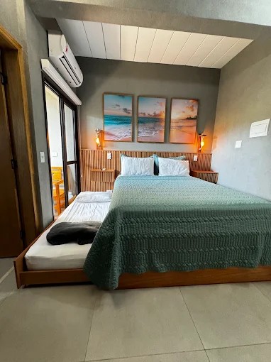 Suíte do Palulu Flat com cama de casal, piso claro e esquadria voltada para a área externa